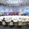 Reunión de la segunda sesión de la cumbre BRICS 2024 en Rusia, con representantes de varios países sentados en una mesa redonda, rodeados de banderas nacionales y bajo una intensa iluminación blanca. En el fondo, se aprecia el logo tricolor del evento. Mayoría mundial