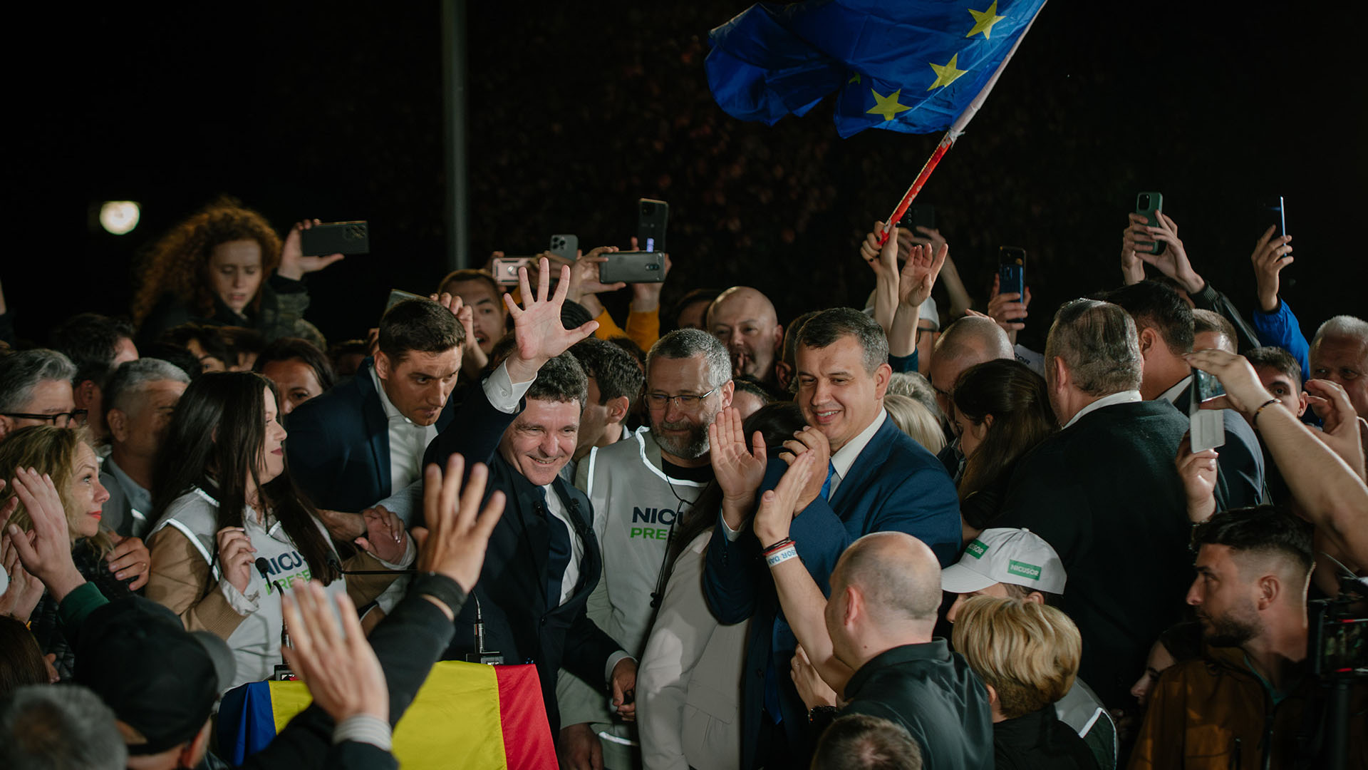 Presidential candidate Nicușor Dan greets his supporters after the first exit poll results during Romania’s presidential runoff election. A European Union flag and a Romanian flag can be seen among the crowd, while several people capture the moment on their mobile phones. Romania
