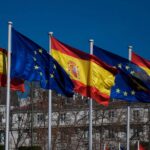 Banderas de España (rojo y amarillo con escudo) y de la Unión Europea (azul con estrellas doradas) ondeando juntas al viento frente a un edificio en un día soleado. Comisión Europea