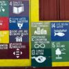 Mural pintado en una pared que representa los 17 Objetivos de Desarrollo Sostenible (ODS) de las Naciones Unidas, cada uno con un ícono y color distintivo. A la izquierda del mural hay una puerta roja con un cartel que dice "NO VENDING". A la derecha, sobre el mural, se encuentra una ventana de madera color terracota, enmarcada por una franja amarilla. Desarrollo