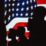 Human silhouettes in front of a brightly lit Unites States flag screen. US