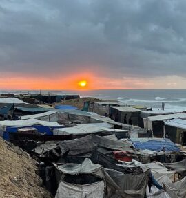 Campamento improvisado de personas desplazadas en la costa de la Franja de Gaza al atardecer, con refugios y tiendas hechas de lonas y materiales diversos, agrupados bajo un cielo nublado. Al fondo se ve el mar y el sol parcialmente oculto tras las nubes grises. Gaza