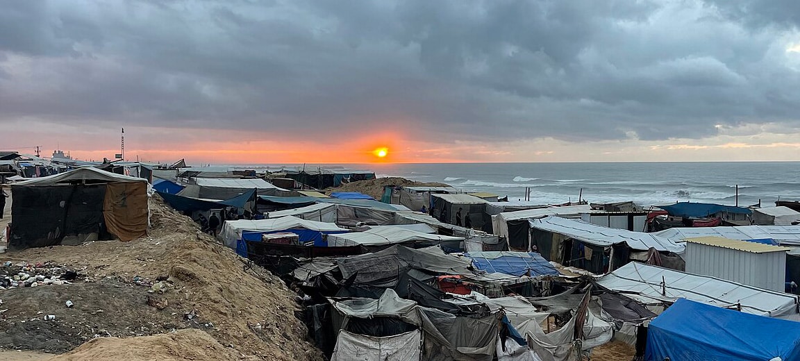 Improvised camp for displaced people on the coast of the Gaza Strip at sunset, with tents and shelters made of tarpaulins and assorted materials, clustered closely under a cloudy sky. In the background, the sea and the sun are partially hidden behind grey clouds. Gaza