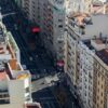 Vista aérea diurna de la Gran Vía en Madrid (España), en un día soleado. A ambos lados de la avenida destacan edificios de diferentes colores y estilos arquitectónicos. En el centro, la calzada y el área peatonal muestran vehículos y peatones desplazándose en diversas direcciones. España
