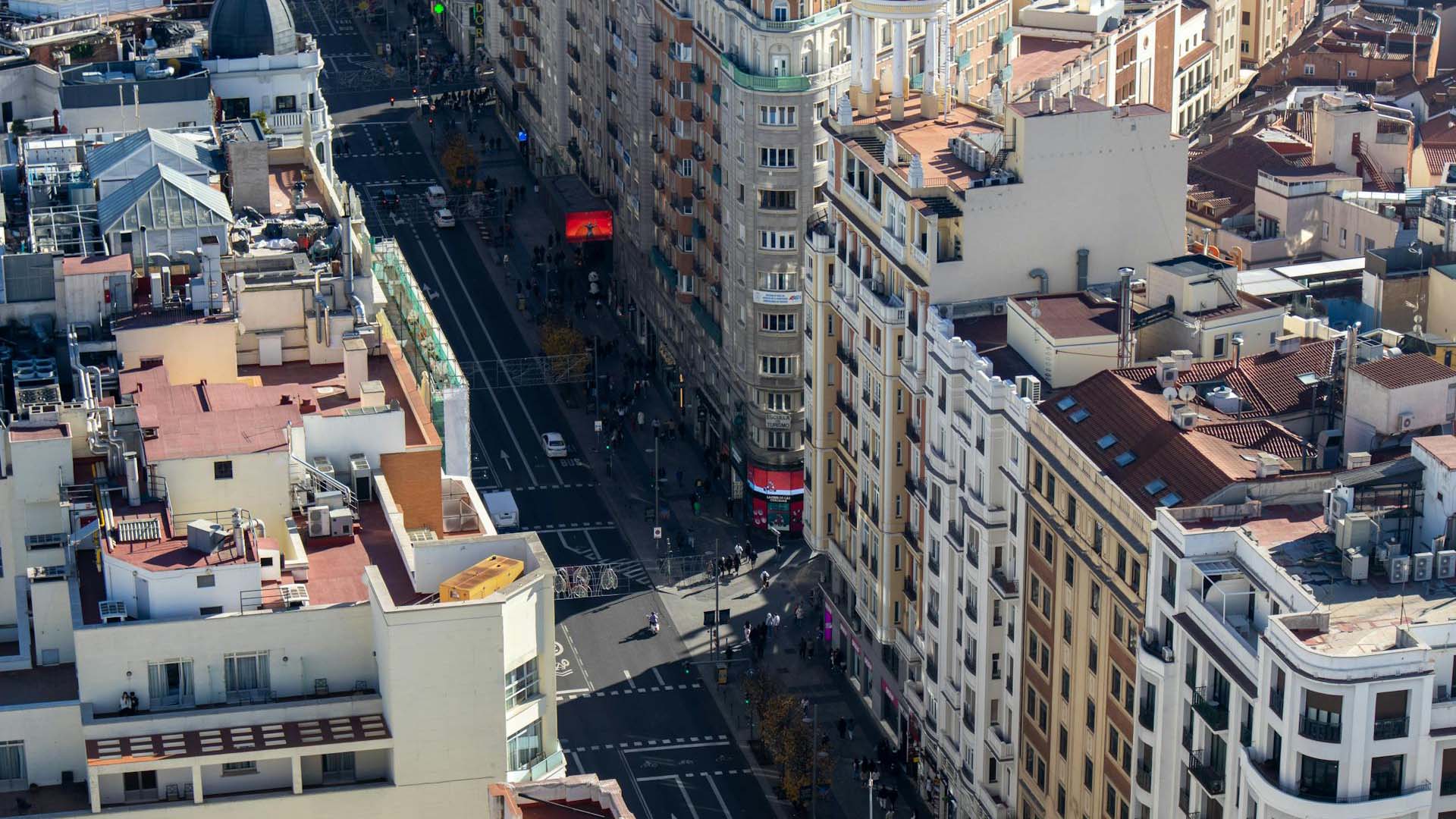 Vista aérea diurna de la Gran Vía en Madrid (España), en un día soleado. A ambos lados de la avenida destacan edificios de diferentes colores y estilos arquitectónicos. En el centro, la calzada y el área peatonal muestran vehículos y peatones desplazándose en diversas direcciones. España