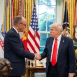 2El presidente Donald Trump saluda al canciller alemán Friedrich Merz con un apretón de manos en el Despacho Oval. Ambos líderes visten trajes oscuros y permanecen de pie frente a un despliegue de banderas estadounidenses y estandartes militares. Un busto de Abraham Lincoln y fotografías enmarcadas son visibles en el fondo, junto con grandes ventanas que proporcionan luz natural. Alemania