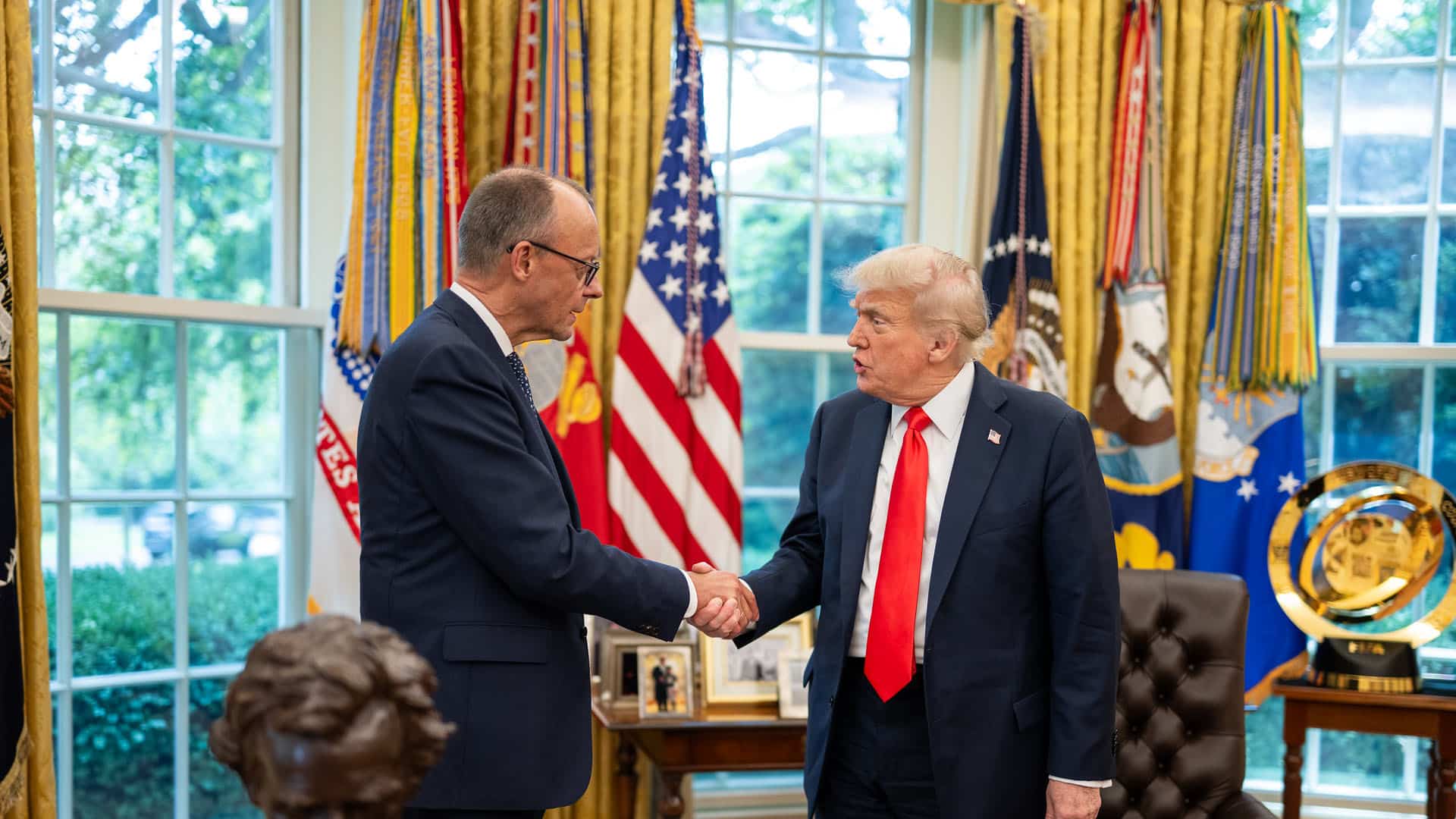 2El presidente Donald Trump saluda al canciller alemán Friedrich Merz con un apretón de manos en el Despacho Oval. Ambos líderes visten trajes oscuros y permanecen de pie frente a un despliegue de banderas estadounidenses y estandartes militares. Un busto de Abraham Lincoln y fotografías enmarcadas son visibles en el fondo, junto con grandes ventanas que proporcionan luz natural. Alemania