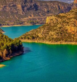 Panorámica del embalse de Camarasa (Cataluña), con aguas esmeralda rodeadas de montañas escarpadas y frondosos bosques, bajo un cielo azul que contrasta con algunas nubes. Agua