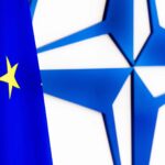Flag of the European Union (EU) in front of the NATO emblem on a white wall.