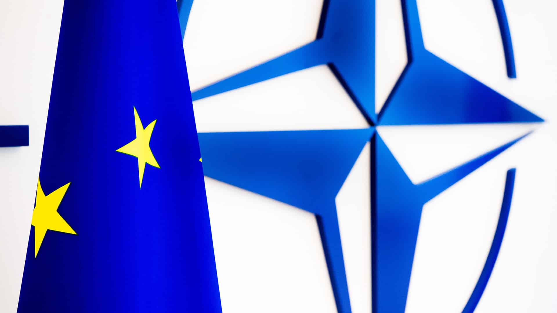 Flag of the European Union (EU) in front of the NATO emblem on a white wall.