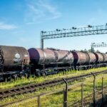 Vagones cisterna con petróleo ruso alineados sobre una vía férrea en una estación cerca de Ivanogrod (Rusia), con una cerca metálica en primer plano y una estructura metálica elevada al fondo. El cielo está parcialmente despejado y hay vegetación verde a los lados de las vías.