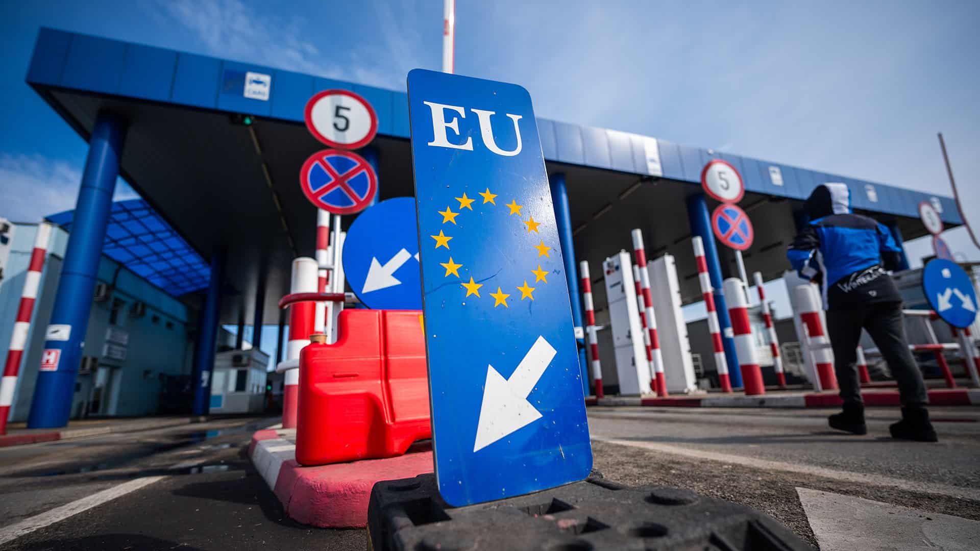 A wide-angle view of the Calafat crossing between Bulgaria and Romania: in the foreground, a blue sign bearing the EU emblem and a white arrow; beyond it, control booths, red-and-white retractable barriers, speed-limit signs, and an attendant in a blue jacket walking away under a clear sky. Schengen