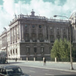 The Royal Palace of Madrid (Spain) in the 1960s. A large, historic building with classical architecture, featuring ornate details and a dome on the right side. The building is situated along a street with several old cars and a double-decker bus. Trees are visible in front of the building, and the sky is partly cloudy.