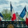 Tres banderas francesas ondean en un puente con barandilla de hierro forjado, frente a la entrada de un edificio histórico (Museo de Louvre en París) con cúpula y árboles a ambos lados del río.