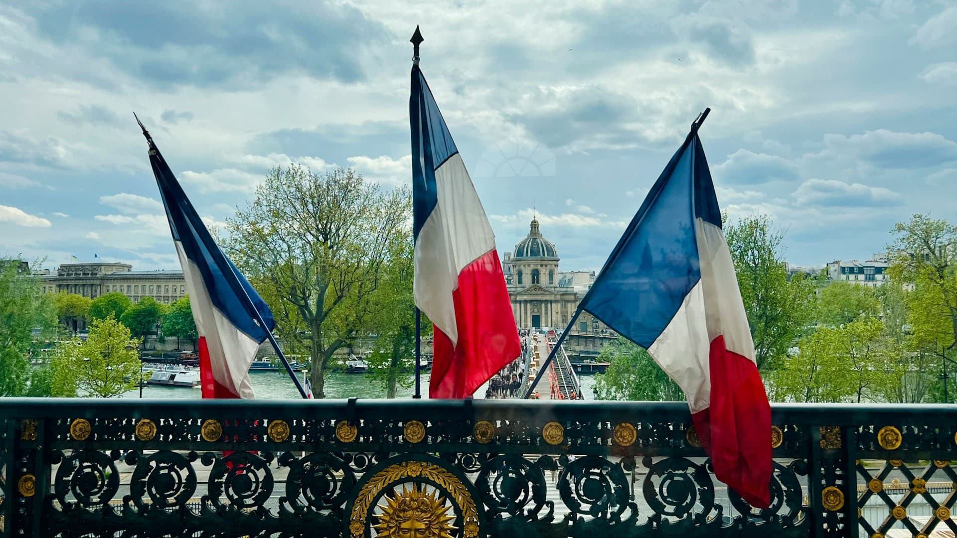 Tres banderas francesas ondean en un puente con barandilla de hierro forjado, frente a la entrada de un edificio histórico (Museo de Louvre en París) con cúpula y árboles a ambos lados del río.