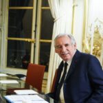 François Bayrou, primer ministro de Francia, sentado en una mesa de reuniones con documentos, portátil y vasos de agua, en una sala elegante con cortinas blancas y detalles dorados con banderas de Francia y la Unión Europea al fondo. Imagen recortada del encuentro con Magnus Brunner, comisario europeo de Asuntos Internos y Migración, en París. (20/01/2025).
