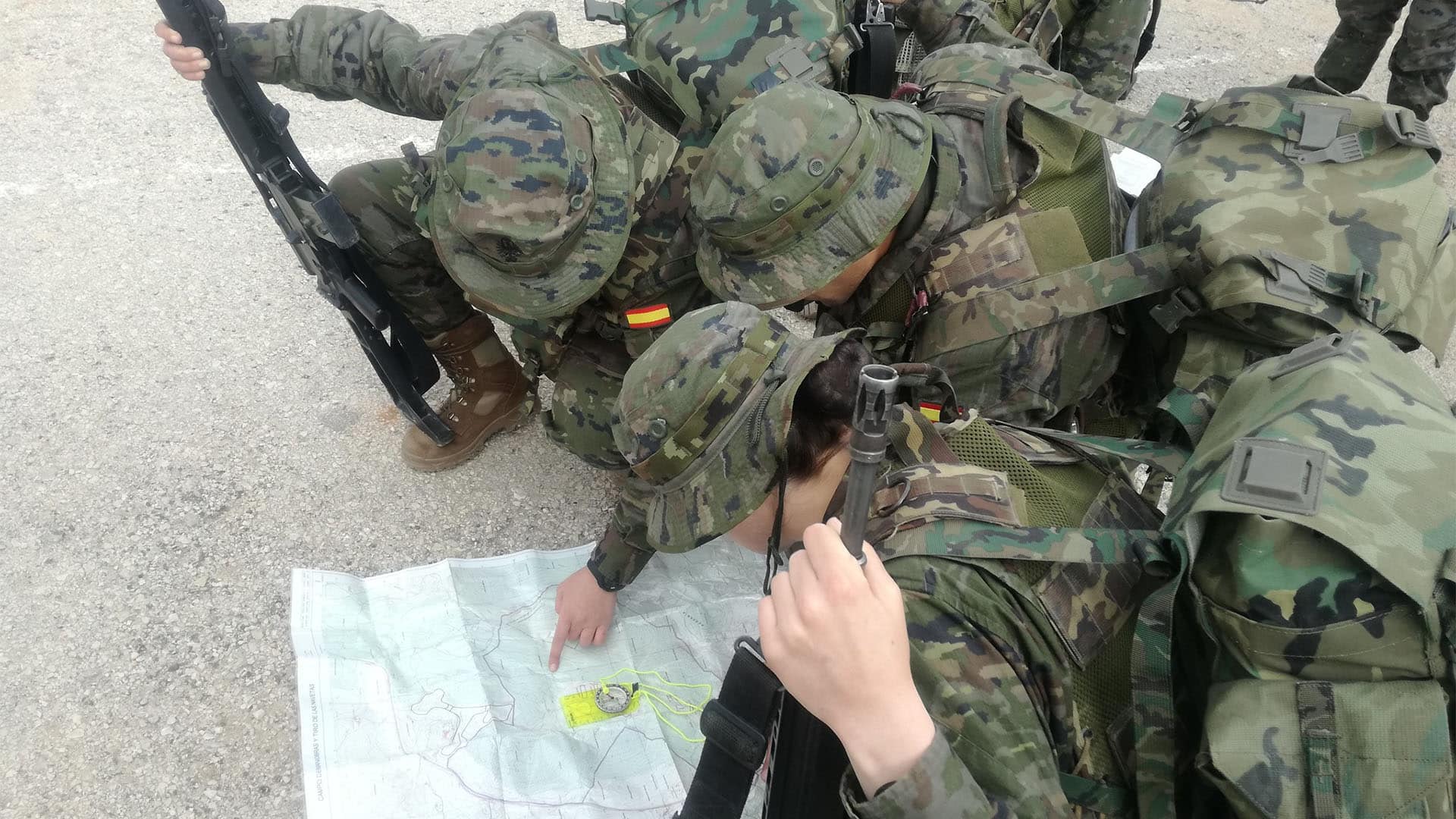 Jóvenes (entre ellos inmigrantes) aspirantes a soldados profesionales del Ejército de Tierra en el Centro de Formación de Tropa nº 2 (2019) con uniformes de camuflaje revisando un mapa desplegado en el suelo; uno de ellos señala una zona del mapa mientras portan mochilas y rifles.