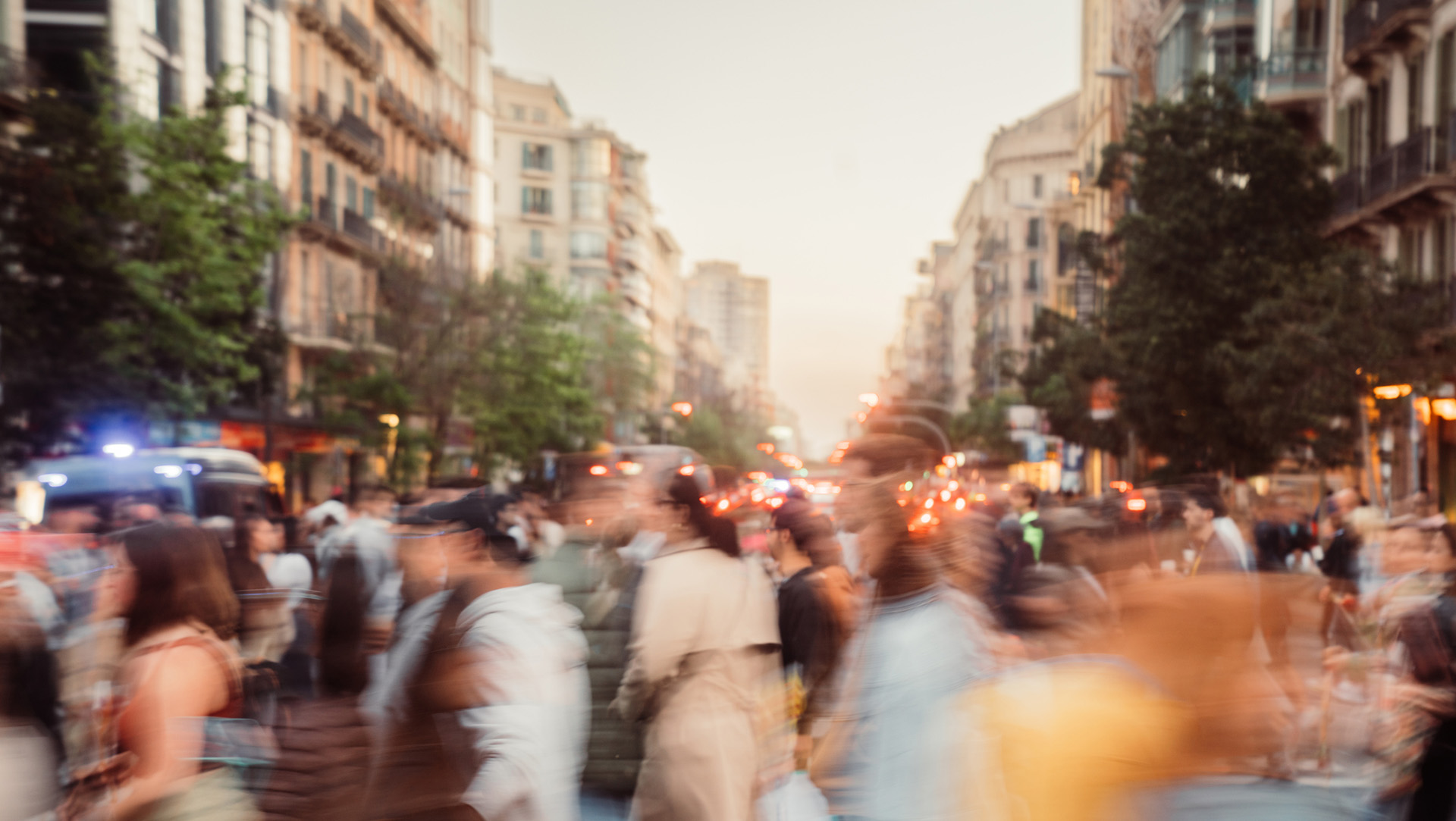 A busy street of Barcelona (Spain) at dusk with blurred motion of people walking. Tall buildings and trees line the street, illuminated by a mix of natural and artificial lighting, creating a lively and bustling atmosphere