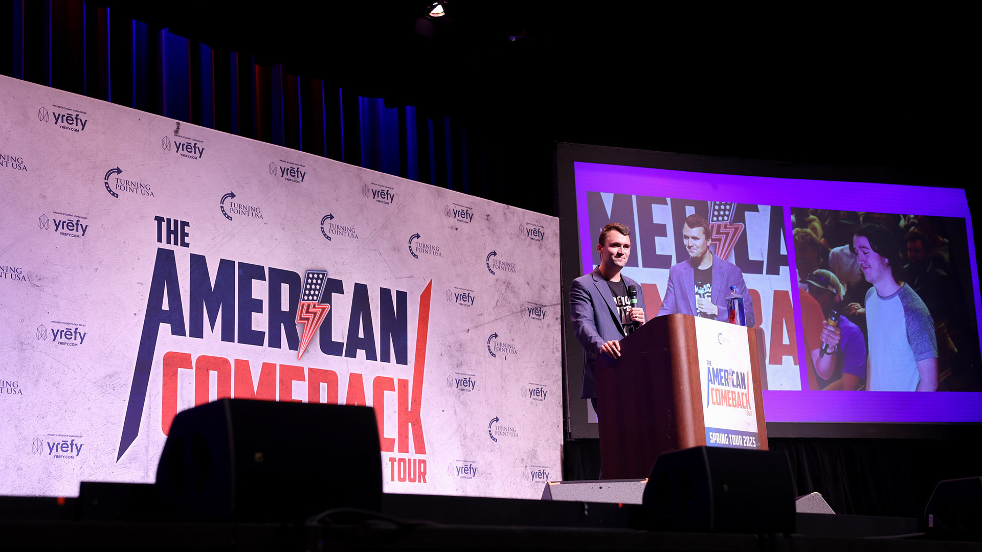 Charlie Kirk responde a preguntas de los asistentes desde el podio del escenario del “American Comeback Tour”, mientras una pantalla ampliada dividida muestra su imagen y la de un asistente del evento. Al fondo, el logo del evento.
