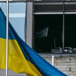 Una bandera de Ucrania ondea en un mástil frente a un edificio dañado por los bombardeos por la guerra de Rusia en Kyiv. Al fondo, cristales rotos, paredes oscurecidas por el fuego y restos de mobiliario de oficina.
