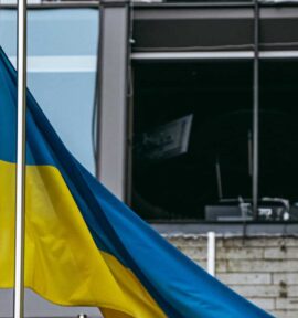 A Ukrainian flag flies from a flagpole in front of a building damaged by bombing during the Russian war in Kyiv. In the background, broken glass, walls blackened by fire and the remains of office furniture.