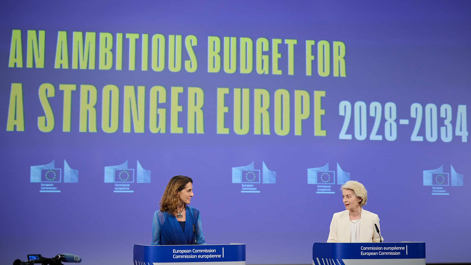 Paula Pinho, portavoz principal de la Comisión Europea (a la izquierda) y Ursula von der Leyen, presidenta de la Comisión Europea (a la derecha) hablando desde los atriles ante la prensa durante el resumen de la reunión semanal de la Comisión sobre el próximo presupuesto de la UE. A la izquierda se asoma una cámara de televisión y al fondo la pantalla azul proyecta el logo de la Comisión en fila y las palabras en inglés “An ambituos budget for a stronger Europe 2028-2034” (un presupuesto ambicioso para una Europa más fuerte 2028-2034).