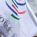 Bandera blanca con el logotipo de la Organización Mundial del Comercio (OMC) en su sede de Ginebra (Suiza). El edificio tiene la palabra «La Paix» en el tejado y una bandera suiza ondeando.