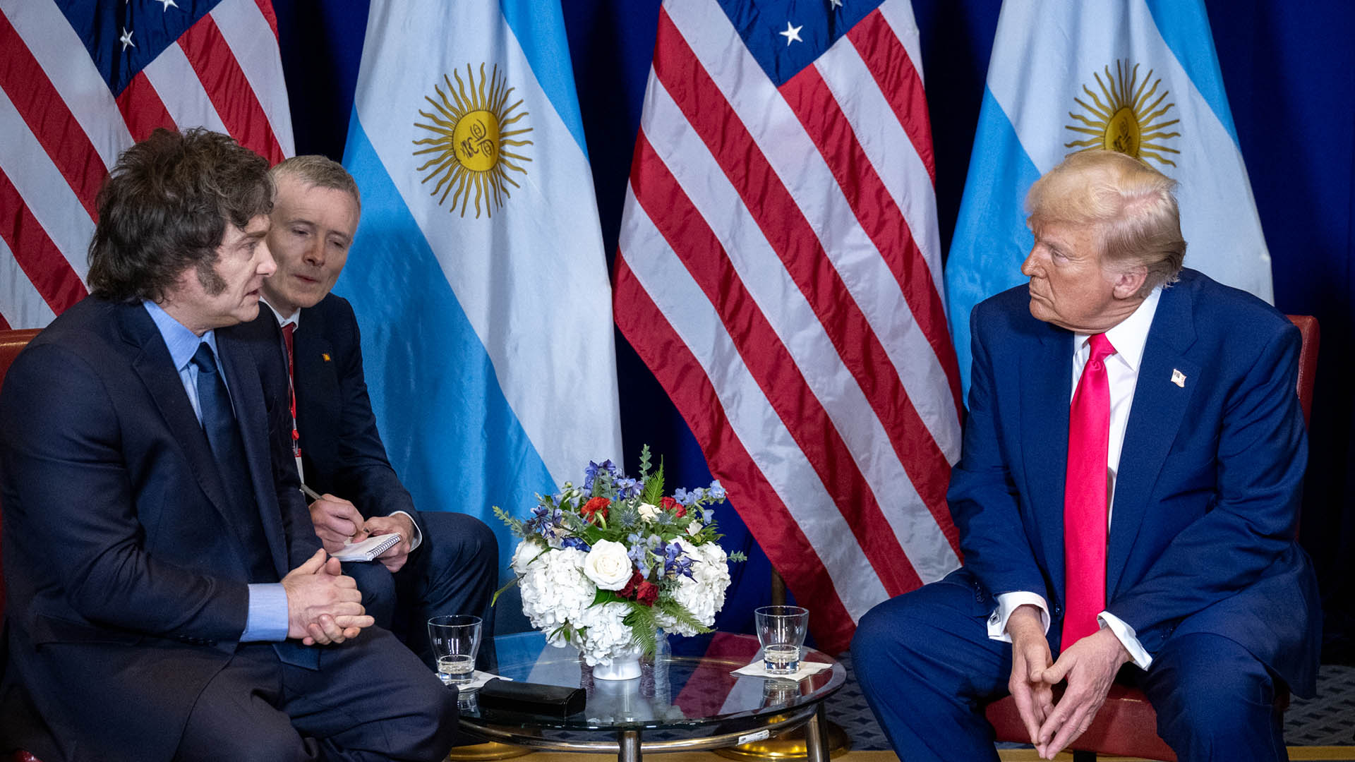 Javier Milei, his translator, and Donald Trump during the meeting between the two leaders at the Gaylord National Resort & Convention Centre in Oxon Hill, Maryland (22/02/2025). In the background, the flags of Argentina and the United States and a floral arrangement on the table in the centre.