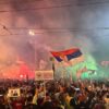 Gran protesta nocturna llamada «Nos vemos en Vidovdan», con multitud de personas portando banderas de Serbia, pancartas y teléfonos móviles, rodeados de humo y luces de colores.