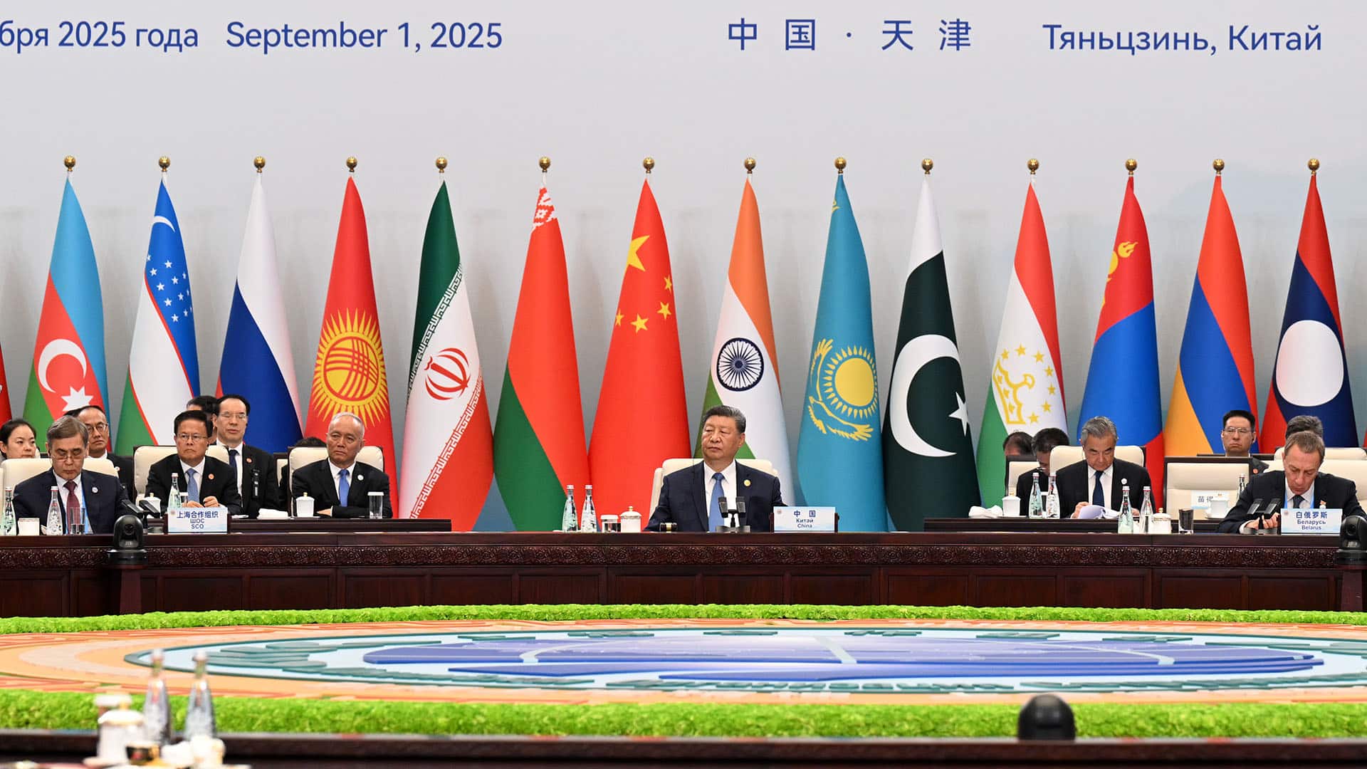 Main table at the plenary session of the 25th Meeting of the Council of Heads of State of the Shanghai Cooperation Organization (SCO) in Tianjin, China. In the centre, Xi Jinping (President of China) flanked by the other representatives and delegations. In the background, the flags of the SCO member countries, with text indicating the date (1 September 2025) and location (Tianjin, China).