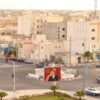 Vista aérea de una intersección en Dajla (ciudad del Sáhara Occidental) con edificios bajos, tráfico moderado y una rotonda central decorada con un gran cartel que muestra un retrato oficial del Rey Mohamed VI de Marruecos, en un entorno urbano desértico.
