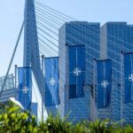 Several vertical NATO blue flags fly in front of a modern cable-stayed bridge and tall glass buildings in an urban setting in Rotterdam, the Netherlands, on the NATO Day in April 2023.