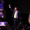 Zohran Mamdani celebra su victoria en el escenario con el brazo en alto durante la fiesta electoral celebrada en el Brooklyn Paramount, en el distrito de Brooklyn de la ciudad de Nueva York (4/11/202). A un lado, la bandera de Estados Unidos en un mástil, y al otro parte de un cartel lila con las palabras “For New York City”.