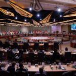 Delegates from the countries attending the 2025 G20 Summit meeting at the Johannesburg Expo Centre (22/11/2025). They are all seated around several concentric circles with geometric shapes and flowers in the centre. On the ceiling, several geometric shapes are illuminated, and in the background, the flags of the participating countries can be seen. There are also several screen panels on both sides of the conference room.