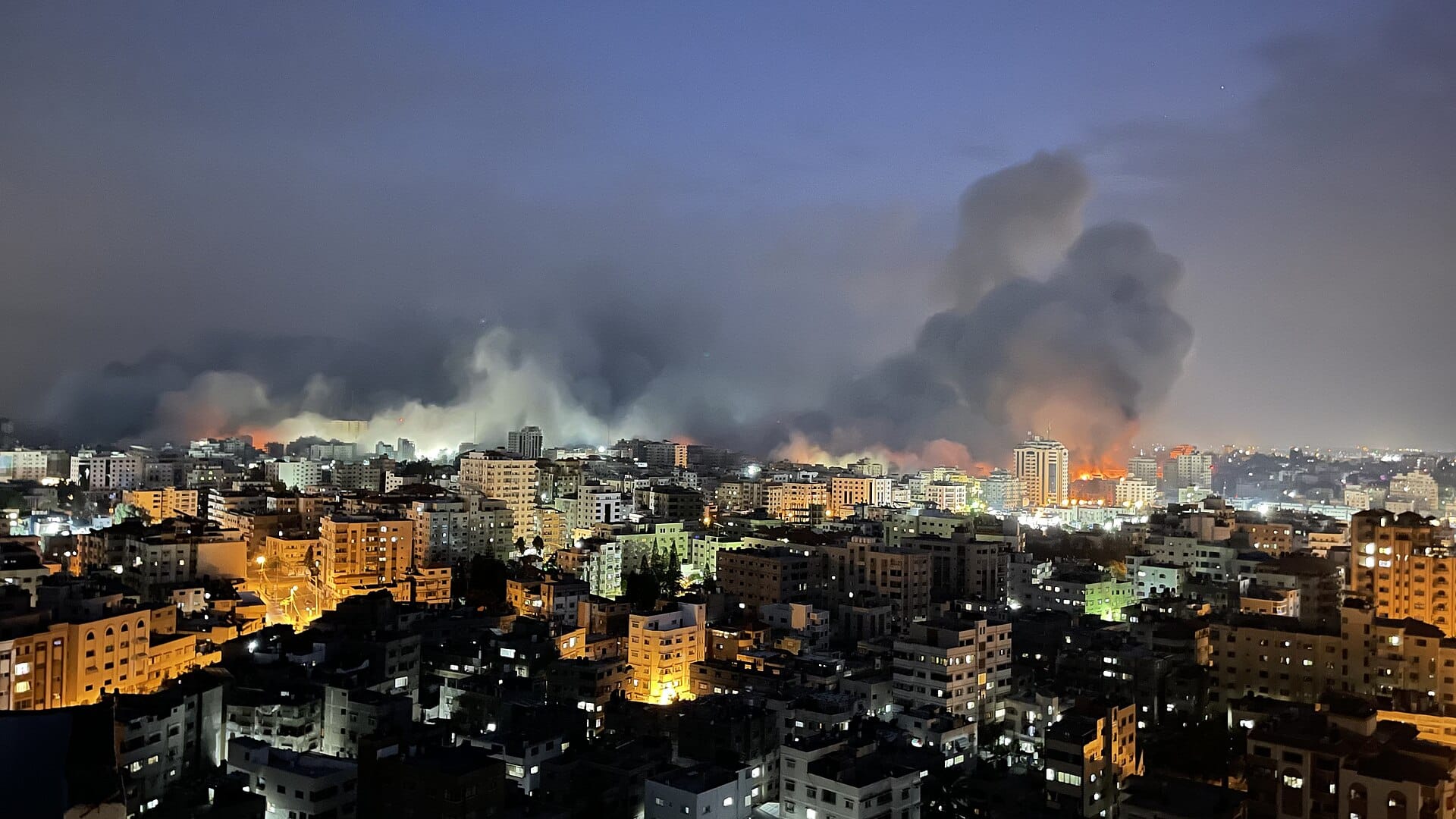 Los ataques aéreos israelíes incendian las calles de la Franja de Gaza durante un bombardeo en febrero de 2025 (10/02/2025). Los focos de los bombardeos incendian e iluminan algunas calles de la ciudad, mientras, en el fondo, una gran nube de humo oculta los edificios del horizonte y el cielo nocturno.