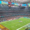 Vista del estadio Santiago Bernabéu durante el partido NFL Miami Dolphins vs Washington Commanders (16/11/2025). El campo está vacío y los jugadores se encuentran agrupados fuera de él, cada equipo a un lado. En el centro del campo, el logo de la NFL y a los lados los logos de ambos equipos. Las gradas están llenas y en la pantalla superior aparece el fondo azul de los Dolphins con el texto “2025 NFL Madrid Game”, la retransmisión en directo y el marcador: Commanders 0 – Dolphins 3. Sobre la pantalla cuelgan las banderas de España, la NFL y Estados Unidos.