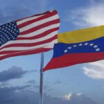 The flags of the United States and Venezuela flying in a clear sky. In the foreground, the flag of the United States, and in the background, the flag of Venezuela, fly above the flagpoles against a clear sky with a few clouds.