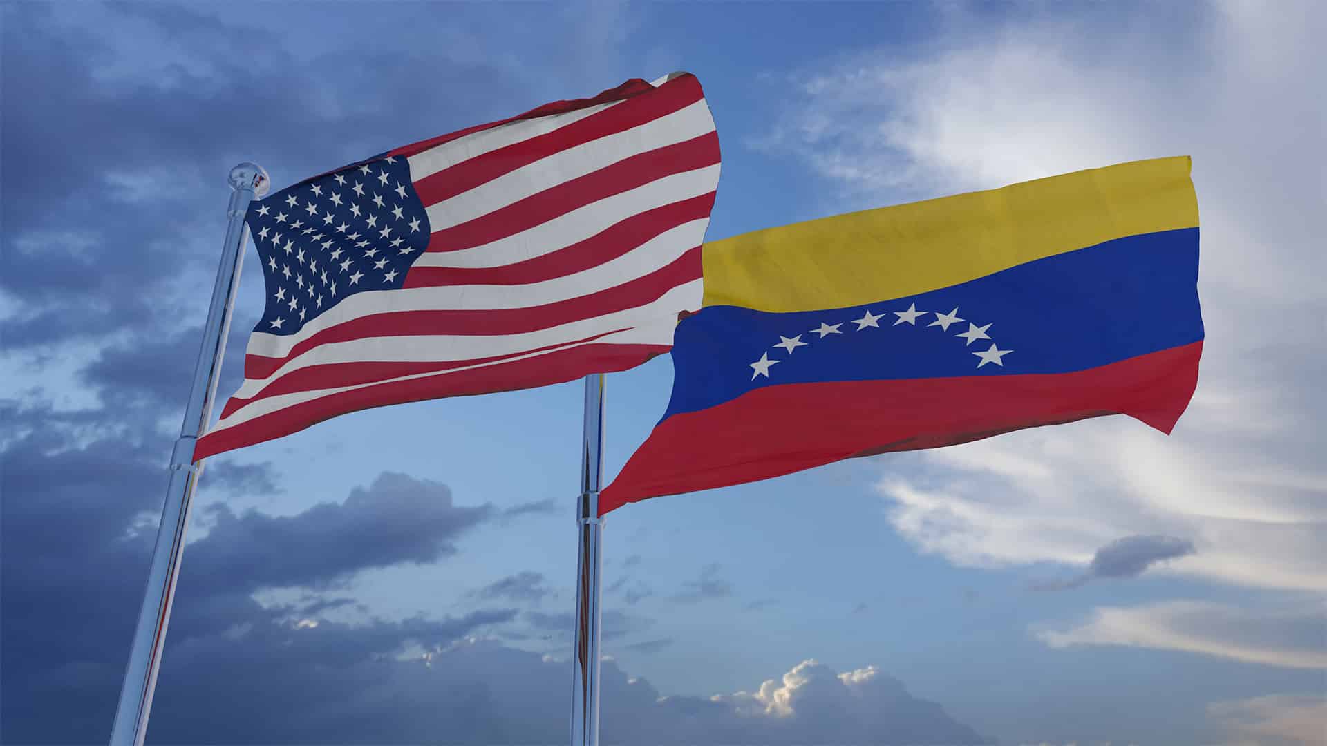 The flags of the United States and Venezuela flying in a clear sky. In the foreground, the flag of the United States, and in the background, the flag of Venezuela, fly above the flagpoles against a clear sky with a few clouds.