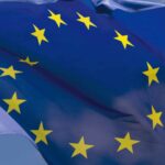 The flag of the European Union flies in the foreground against a clear, cloudless sky.