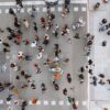 Vista superior de un cruce peatonal muy concurrido en España. A ambos lados, dos aceras amplias donde los transeúntes esperan para cruzar en el semáforo. En el centro, el paso de peatones está abarrotado de personas que avanzan en ambas direcciones. En la parte superior, hay vehículos detenidos antes del semáforo, mientras que en la inferior aparece parte de los semáforos que regulan el cruce.