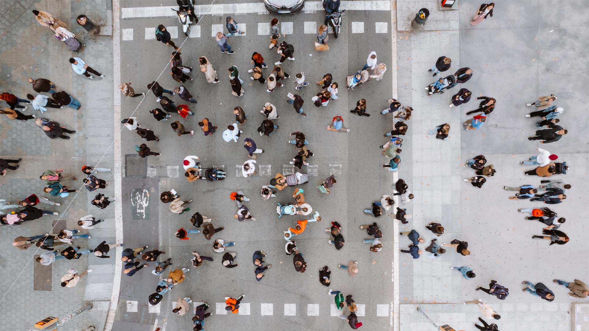Vista superior de un cruce peatonal muy concurrido en España. A ambos lados, dos aceras amplias donde los transeúntes esperan para cruzar en el semáforo. En el centro, el paso de peatones está abarrotado de personas que avanzan en ambas direcciones. En la parte superior, hay vehículos detenidos antes del semáforo, mientras que en la inferior aparece parte de los semáforos que regulan el cruce.