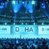 Large conference room at the Doha Forum with attendees in front of a blue-lit stage and two screens displaying the word ‘DOHA’.
