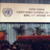 Hillary Clinton pronuncia su discurso en la IV Conferencia Mundial sobre la Mujer (Beijing, 5 de septiembre de 1995) en el escenario del Beijing International Conference Center. Viste un traje rosa y se dirige al público desde una tribuna adornada con flores y el logo de la ONU. A la derecha, se observa otra mesa con representantes y micrófonos, también decorada. Al fondo, un telón azul, un gran cartel con el texto: “United Nations Fourth World Conference on Women. Beijing, 4-15 September 1995” en la parte superior y, debajo, algunas palmeras.