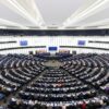 Hemiciclo del Parlamento Europeo en Estrasburgo. Siete filas de mesas con los representantes del parlamento europeo atienden a la asamblea, frente a la mesa de dirección. Detrás, pueden verse las banderas de los estados miembros y la de la Unión Europea, y dos pantallas. Tanto las paredes del hemiciclo como la cubierta, son de color blanco.