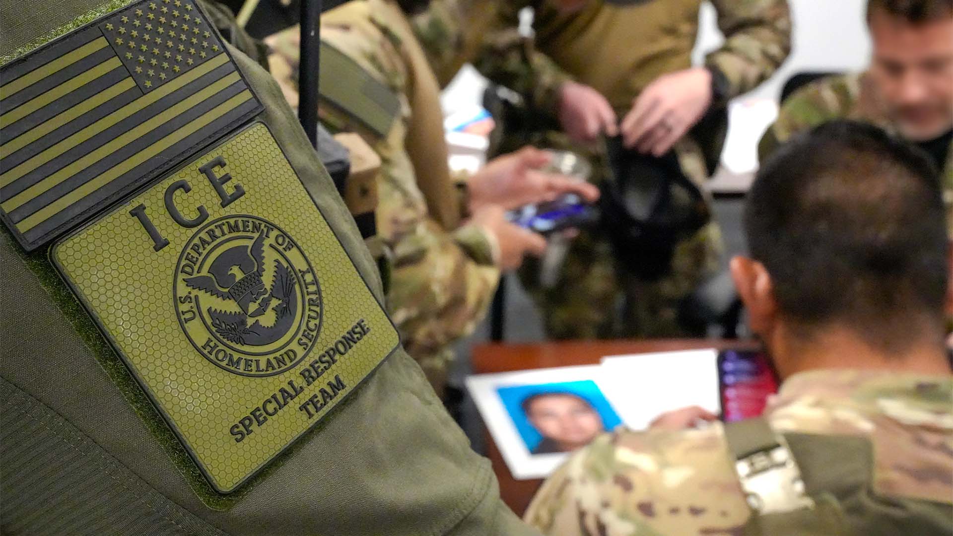 ICE Special Response Team at a briefing prior to an arrest in Rhode Island. In the foreground, part of one of the members' uniforms with the ICE - Special Response Team insignia. Behind, several members of the team in military uniforms gathered around a table with a dossier on top containing a blurred photo of the person they are going to arrest.