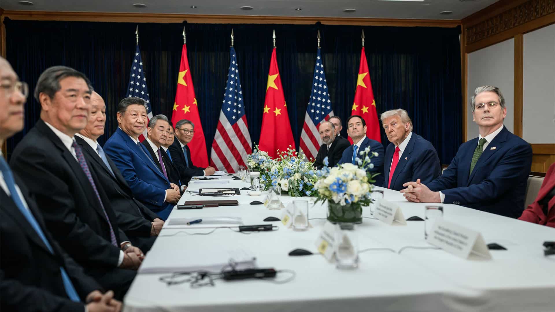 Reunión bilateral entre Donald Trump y Xi Jinping (30/10/2025) en el aeropuerto internacional de Gimhae, en Busán, Corea del Sur. A la izquierda, los representantes de la administración de China, liderada por Xi Jinping; y, a la derecha, la de EEUU, encabezada por Donald Trump, miran hacia la cámara que toma la fotografía. Al fondo, tres banderas de EEUU y tres de China, intercaladas.