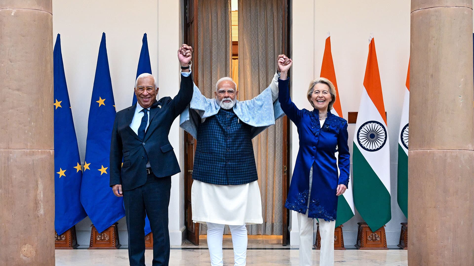 From left to right: António Costa, President of the European Council; Narendra Modi, Prime Minister of India; and Ursula von der Leyen, President of the European Commission, at Hyderabad House, New Delhi, India (27/01/2026). In a gesture of victory, the three representatives raise their hands. On either side, flags of the European Union on the left and India on the right.