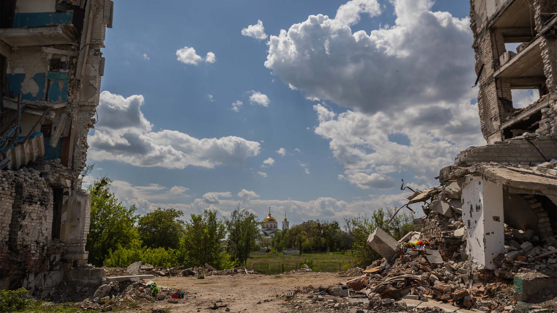 Bloque residencial partido por la mitad debido a los bombardeos en Kyiv. Al fondo, tras un terreno con vegetación y árboles, la cúpula y una torre doradas y blancas de una iglesia, se mantiene intacta. El cielo es claro, aunque con varias nubes.