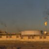 Una planta de refinería de petróleo y una empresa de gas natural en el Golfo Pérsico, Irán. La refinería, de color blanco y delimitada por un muro de ladrillos también blanco, tiene varias llamas que humean en el aire. Al fondo, un paisaje montañoso y un cielo turbio.