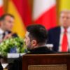 En primer plano, Volodímir Zelenski, presidente de Ucrania, enfatiza con su mano mientras habla. Detrás de él, desenfocados, Emmanuel Macron, presidente de la República Francesa, a la izquierda, y Donald Trump, presidente de EEUU, a la derecha. Los representantes se reúnen en la Casa Blanca, en un encuentro multilateral organizado por el presidente Trump con los líderes europeos.